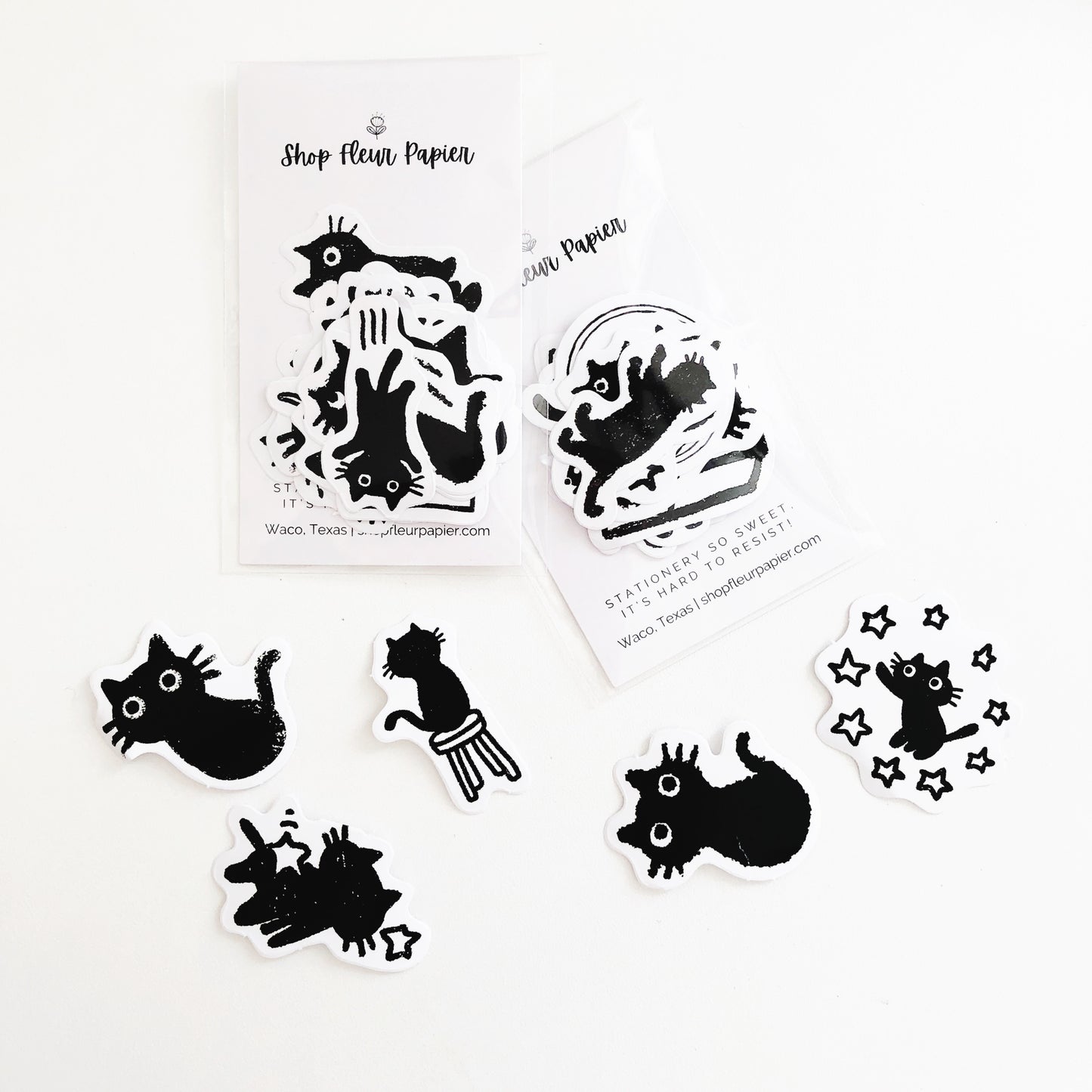 A photo showing a variety of black cat stickers from Shop Fleur Papier. The stickers feature different cat poses and expressions, including a cat playing with a star and a cat sitting on a chair.
