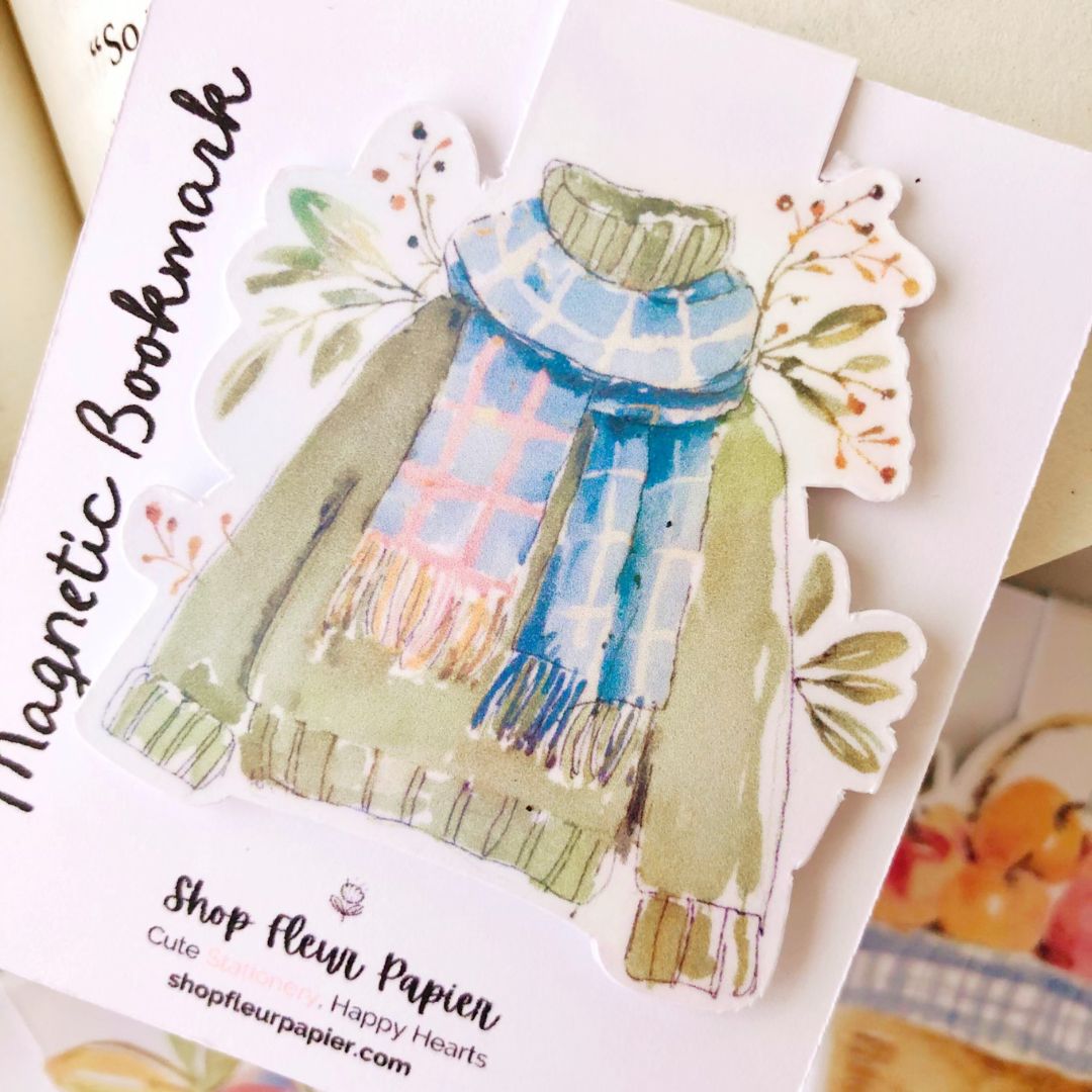 Magnetic bookmark with a design of a green sweater and plaid scarf on a white card.