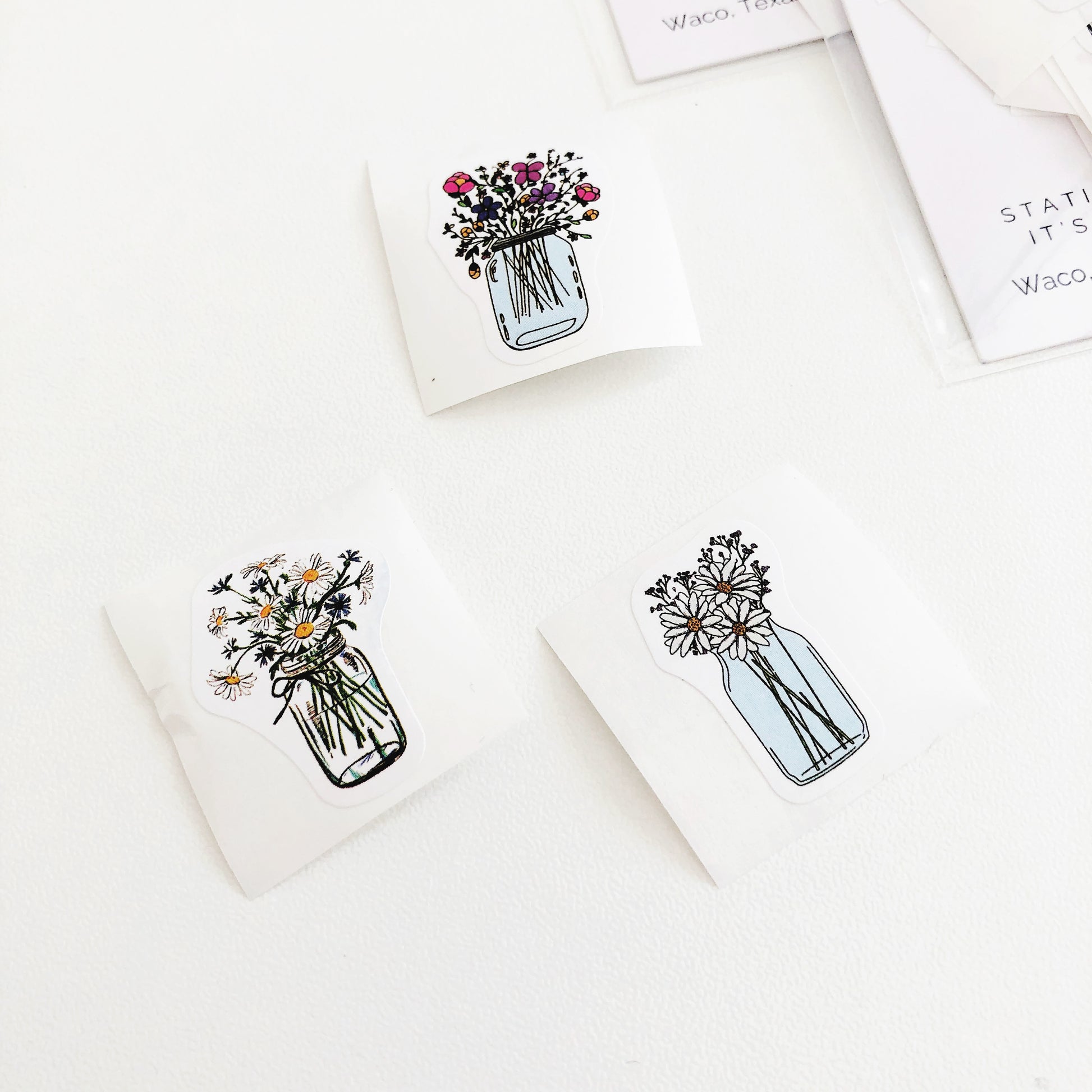 A photo showing three flower stickers from Shop Fleur Papier. The stickers feature different bouquets of flowers in mason jars, with one bouquet featuring colorful flowers and the other two featuring white daisies.