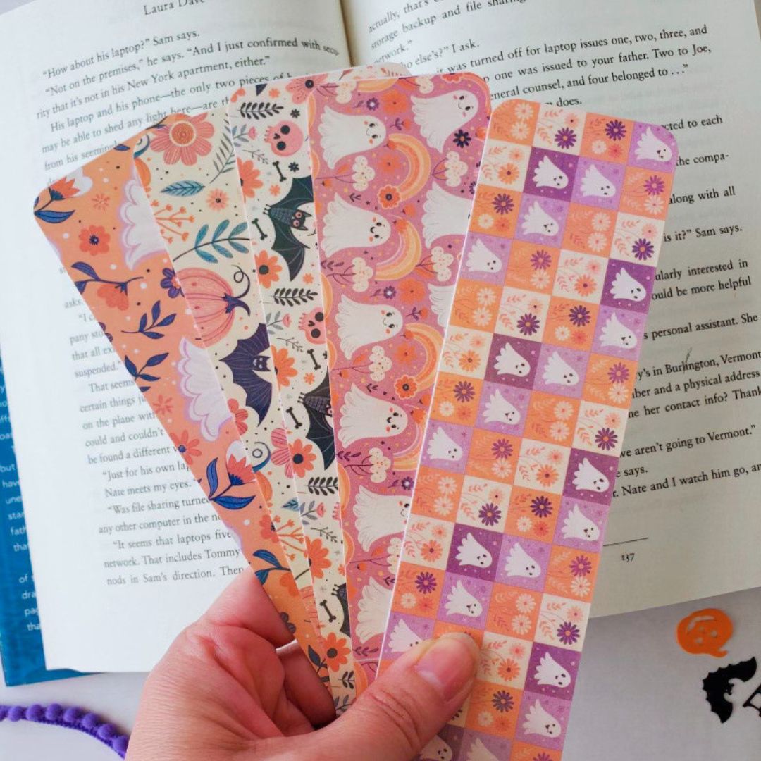 Hand holding a set of five pastel Halloween bookmarks featuring cute ghosts, pumpkins, bats, and floral patterns, styled over an open book.